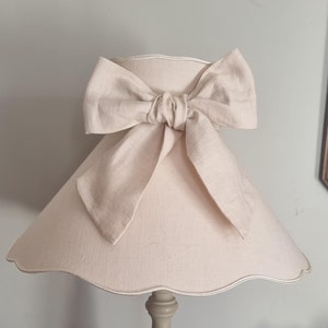 Puede incluir: Una lámpara beige con una pantalla festoneada y un gran lazo. La lámpara tiene una base gris claro con ranuras verticales. La pantalla es de color beige claro.