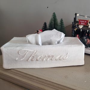 Puede incluir: Una funda blanca rectangular para caja de pañuelos con el nombre "Thomas" bordado en cursiva. La funda está sobre una superficie clara, con una escena de pueblo navideño y árboles en miniatura en el fondo.