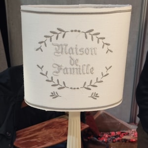 May include: Cream-colored table lamp with a cylindrical lampshade. The lampshade features the embroidered text "Maison de Famille" within a laurel wreath design. The base is square and the stem is fluted.