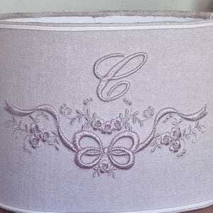K&ouml;nnte beinhalten: Hellgrauer Lampenschirm mit einem gestickten Design. Das Design umfasst einen gro&szlig;en Buchstaben "C", eine Schleife, Blumen und B&auml;nder in passendem Hellviolett. Die oberen und unteren Kanten sind mit einem wei&szlig;en Rand versehen.