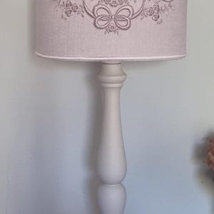 K&ouml;nnte beinhalten: Eine cremefarbene Tischlampe mit Stoffschirm. Der Lampenschirm hat ein gesticktes Design aus einer Schleife und floralen Elementen. Die Lampe hat einen dekorativen Sockel und einen bauchigen Stiel. Die Lampe steht auf einer Holzoberfl&auml;che.