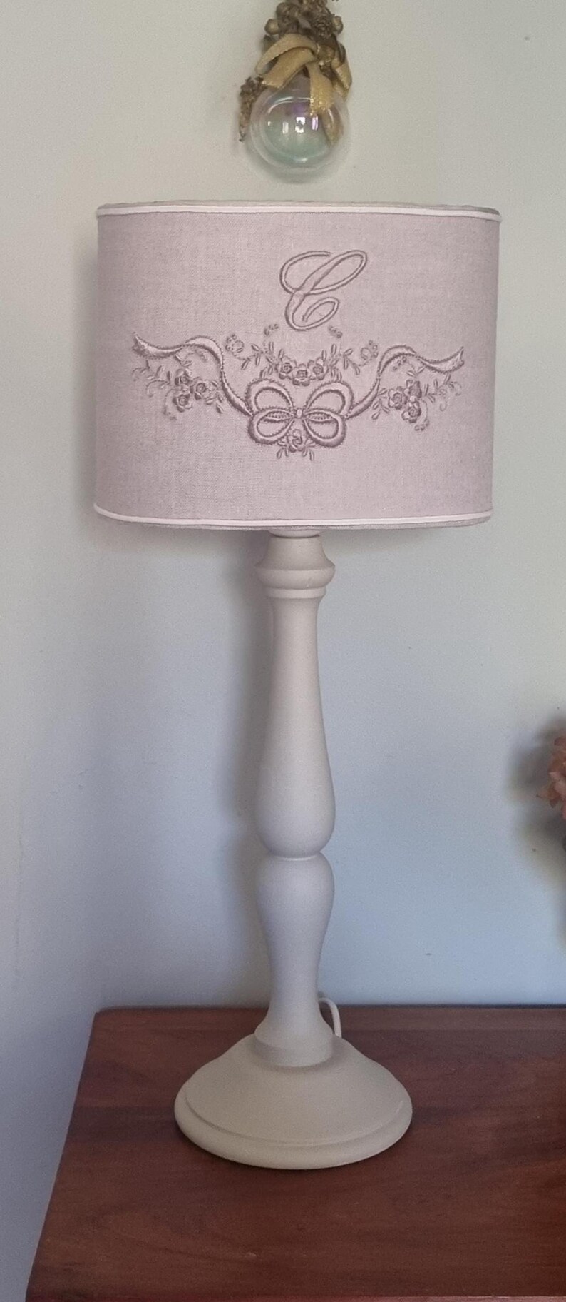K&ouml;nnte beinhalten: Eine cremefarbene Tischlampe mit Stoffschirm. Der Lampenschirm hat ein gesticktes Design aus einer Schleife und floralen Elementen. Die Lampe hat einen dekorativen Sockel und einen bauchigen Stiel. Die Lampe steht auf einer Holzoberfl&auml;che.