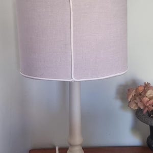 K&ouml;nnte beinhalten: Eine beige Tischlampe mit rundem Sockel und zylindrischem Schirm. Der Schirm hat eine hellviolette Farbe mit wei&szlig;er Umrandung. Die Lampe steht auf einem Holztisch.