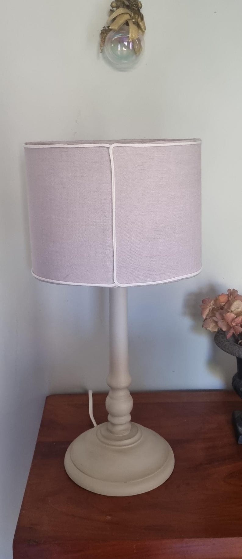 K&ouml;nnte beinhalten: Eine beige Tischlampe mit rundem Sockel und zylindrischem Schirm. Der Schirm hat eine hellviolette Farbe mit wei&szlig;er Umrandung. Die Lampe steht auf einem Holztisch.