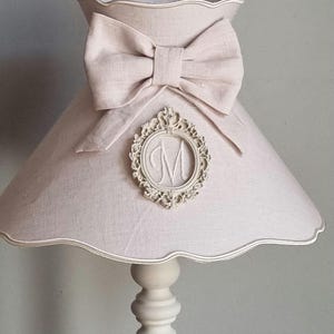 Könnte beinhalten: Eine weiße Lampe mit einem rosa Leinenlampenschirm. Der Lampenschirm hat einen gewellten Rand und eine große Schleife aus dem gleichen Stoff. Ein dekorativer Rahmen mit dem Buchstaben "M" befindet sich mittig auf der Schleife.