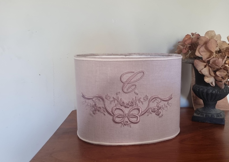 K&ouml;nnte beinhalten: Helllila ovale Lampenschirm mit einem gestickten Design. Das Design umfasst einen gro&szlig;en Buchstaben "C", B&auml;nder und florale Elemente. Der Lampenschirm ist etwa 25 cm hoch.