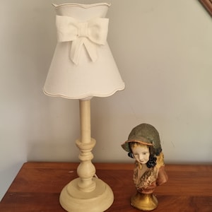 Könnte beinhalten: Eine cremefarbene Tischlampe mit einem schirmförmigen Bogen. Die Lampe hat einen gewellten Rand und einen hellen Holzsockel. Neben der Lampe befindet sich eine dekorative Büste eines Mädchens mit Hut.