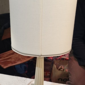 May include: A cream-colored table lamp with a cylindrical shade. The shade is made of a textured fabric and trimmed with a thin gray border. The base is a square shape with a fluted column.