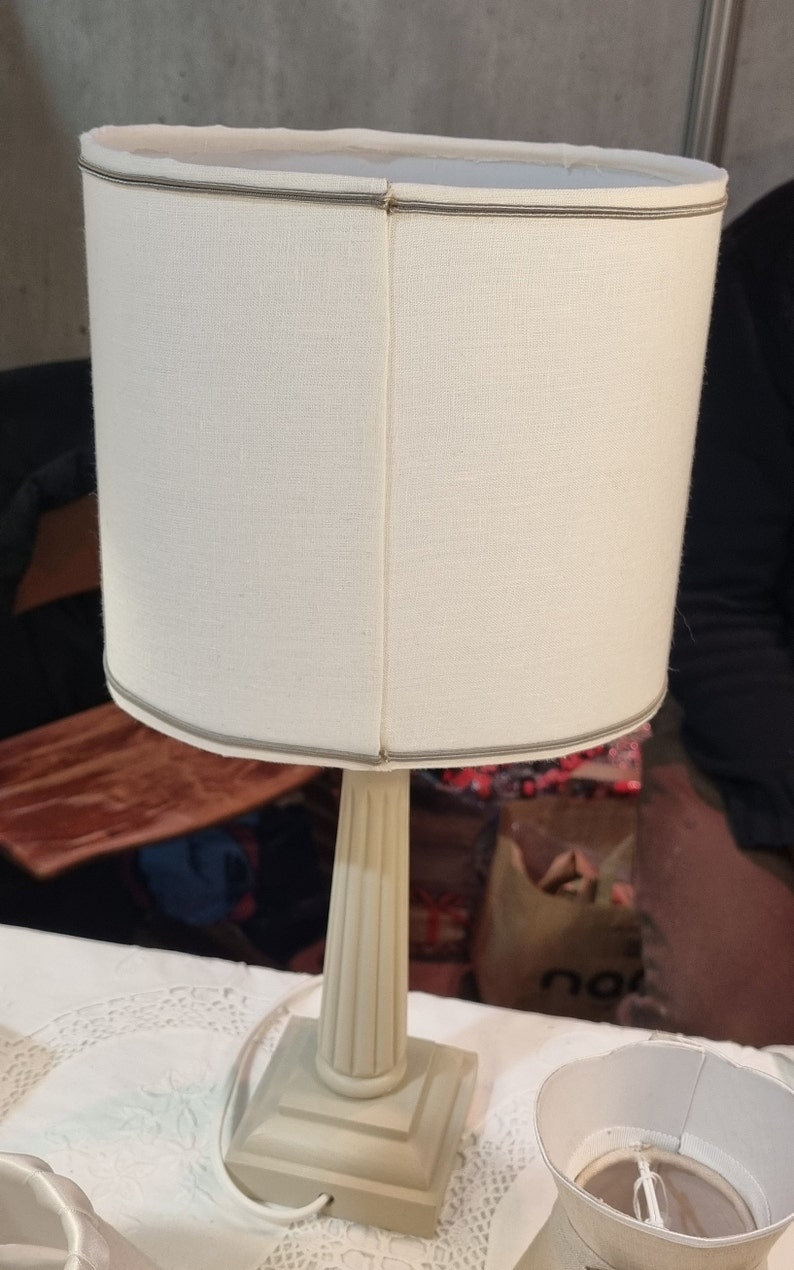 May include: A cream-colored table lamp with a cylindrical shade. The shade is made of a textured fabric and trimmed with a thin gray border. The base is a square shape with a fluted column.