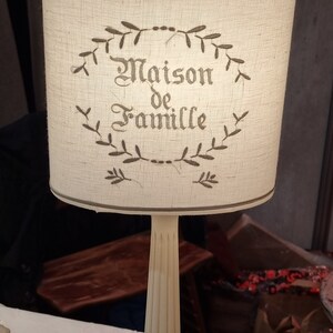 May include: A cream-colored table lamp with a cylindrical lampshade. The lampshade features the text "Maison de Famille" in a decorative font, surrounded by a leafy wreath design. The lamp has a fluted column base and a square base.