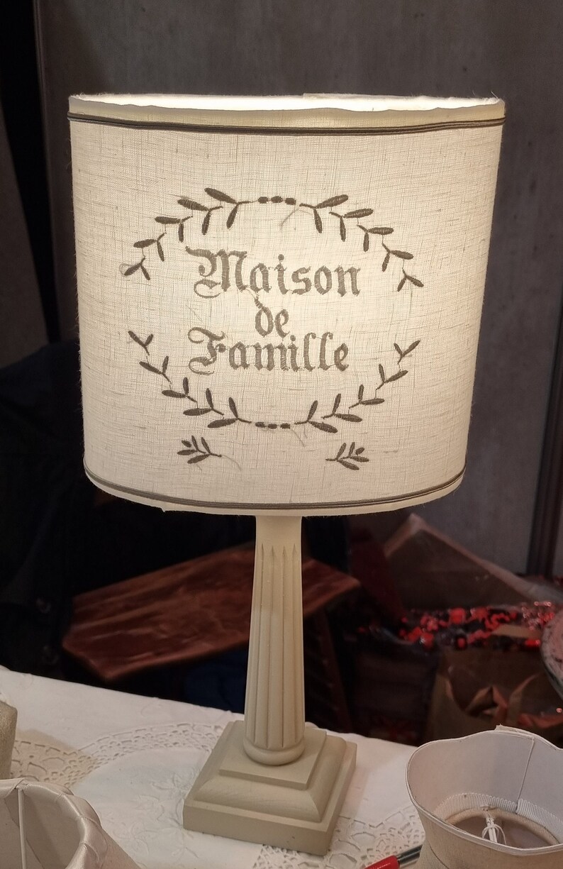 May include: A cream-colored table lamp with a cylindrical lampshade. The lampshade features the text "Maison de Famille" in a decorative font, surrounded by a leafy wreath design. The lamp has a fluted column base and a square base.