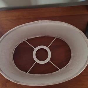 K&ouml;nnte beinhalten: Ovaler Lampenschirm mit cremefarbenem Stoff au&szlig;en. Im Inneren befindet sich ein wei&szlig;es Metallgestell. Der Schirm steht auf einer dunkelbraunen Holzoberfl&auml;che. L&auml;nge ca. 30 cm.