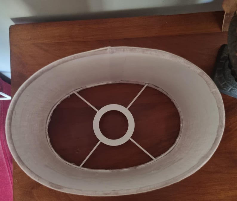 K&ouml;nnte beinhalten: Ovaler Lampenschirm mit cremefarbenem Stoff au&szlig;en. Im Inneren befindet sich ein wei&szlig;es Metallgestell. Der Schirm steht auf einer dunkelbraunen Holzoberfl&auml;che. L&auml;nge ca. 30 cm.