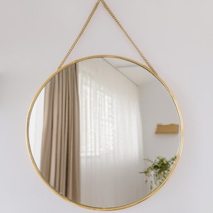 May include: A round gold-framed mirror with a chain hanging from the top. The mirror reflects a room with white curtains and a brown curtain.