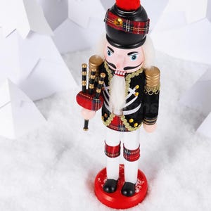 May include: A nutcracker figurine dressed in a black jacket with gold trim, a red and green tartan kilt, and a black hat with a red pom-pom. The figure holds a set of bagpipes and features a white beard, white trousers, and a round red base.