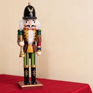 May include: A wooden nutcracker figurine wearing a black helmet with gold wheat detailing, a red and green uniform with gold trim, and white faux fur hair and beard. The figure holds a gold trumpet and stands on a rectangular wooden base.