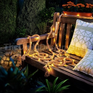 May include: A wooden garden bench adorned with a rope light and two decorative cushions. The rope light, with warm-toned bulbs, is draped across the bench. The cushions are white with green geometric patterns.