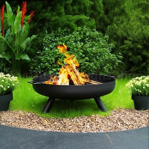 May include: A black metal fire pit with three legs, sitting on a bed of gravel in a grassy garden. The fire pit is filled with burning wood and flames.