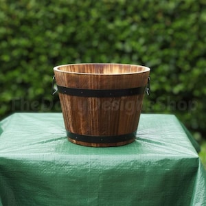 May include: A wooden barrel-style planter with dark metal bands and handles. The planter has a natural wood grain finish and is set on a green surface. The text "The Daily Designs Shop" is visible.