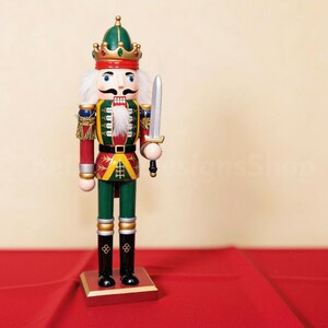 May include: A wooden nutcracker figurine dressed in a green, red, and gold uniform with a matching crown. The figure features a white faux fur beard, holds a silver-coloured sword in its right hand, and stands on a rectangular wooden base.