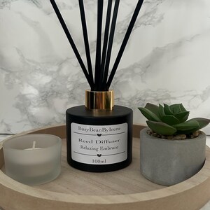 May include: A black reed diffuser with gold detailing and black reeds, labelled "Relaxing Embrace" by BusyBeanByIrene. A small succulent in a grey pot and a candle sit on a wooden tray. The diffuser contains 100ml of liquid.