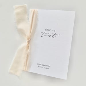 Könnte beinhalten: Eine weiße Karte mit dem Text "Maddie's Toast" und "Maid of Honor August 22, 2026" in schwarzer Schrift. Die Karte ist mit einem cremefarbenen Band mit ausgefransten Kanten gebunden.