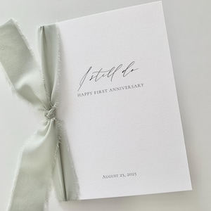May include: A white card with the text "I still do" and "Happy First Anniversary" in elegant script. The date "August 23, 2025" is printed at the bottom. A light green ribbon is tied around the card.