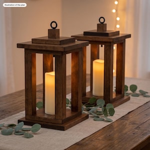 May include: Two square wooden lanterns with a dark brown finish, each featuring a black metal ring on top and a white pillar candle inside. The lanterns have an open-frame design with four vertical posts and a flat base. Text reads: Illustration of the plan.