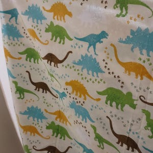 May include: White fabric with a repeating pattern of colorful dinosaurs in shades of green, blue, yellow, and brown. Dinosaur footprints are scattered throughout the design. The dinosaurs are various species.
