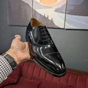 May include: A black leather Oxford shoe with a glossy finish. The shoe features a cap toe design and black laces. The shoe is held up in front of a painting of a mountain landscape.