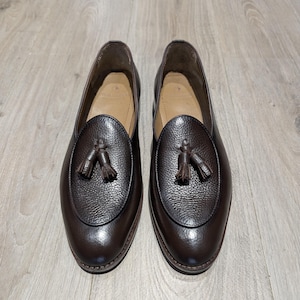May include: A pair of brown leather loafers with a textured finish. Each shoe features a decorative tassel and a rounded toe. The shoes are displayed on a light-colored wooden surface, showcasing their polished appearance.