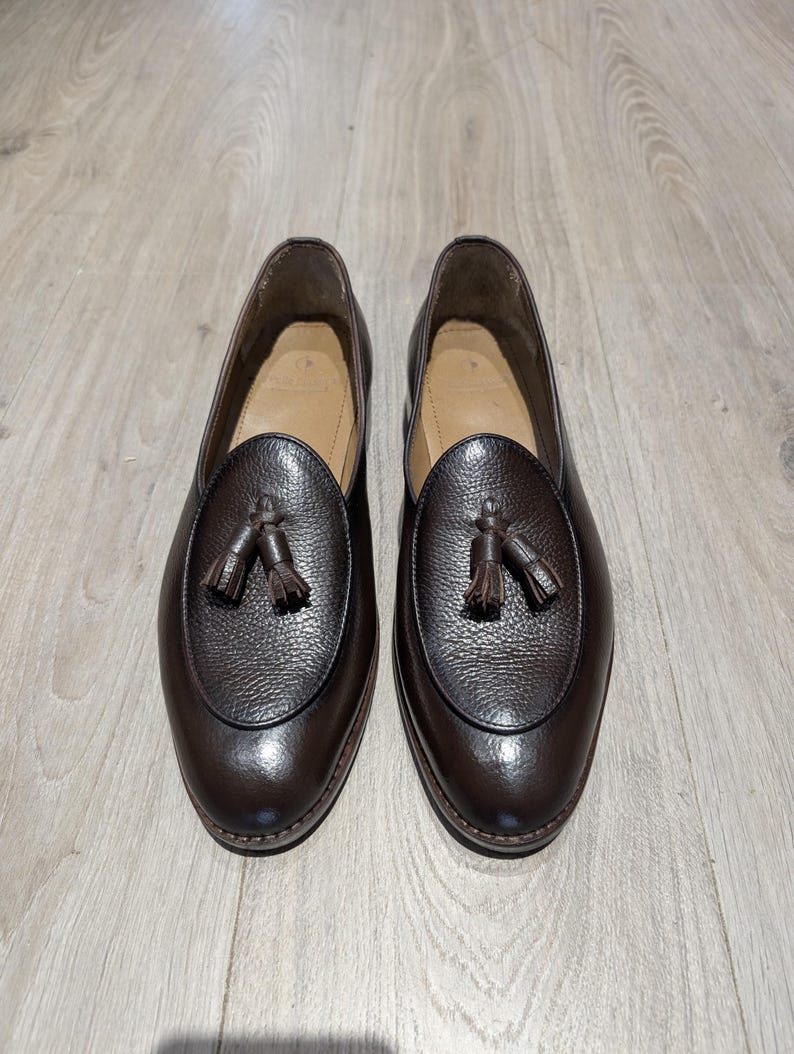 May include: A pair of brown leather loafers with a textured finish. Each shoe features a decorative tassel and a rounded toe. The shoes are displayed on a light-colored wooden surface, showcasing their polished appearance.
