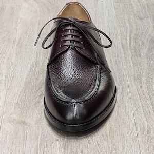 Peut inclure: Une chaussure en cuir marron avec une surface texturée et un bout arrondi. La chaussure a une fermeture à lacets et est posée sur un fond clair en grain de bois. La semelle de la chaussure est noire.