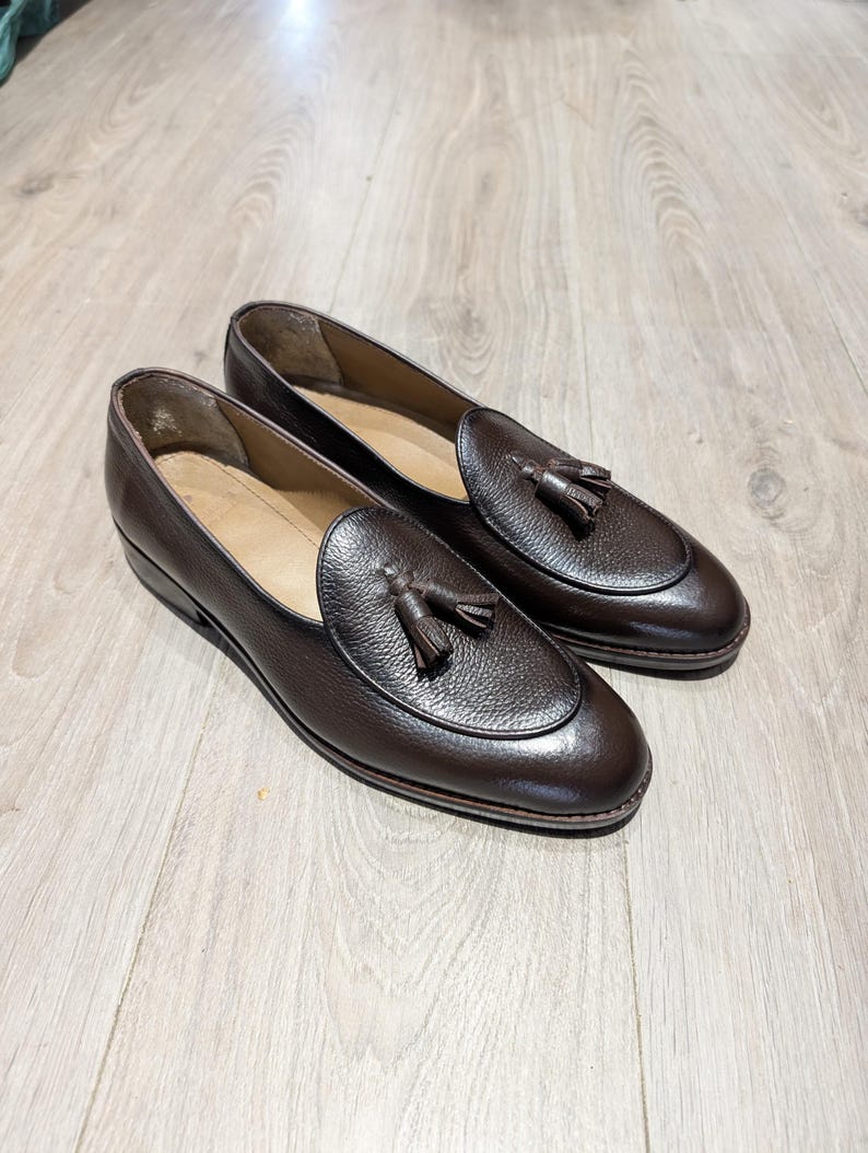 May include: A pair of brown leather loafers with tassel details. The shoes have a rounded toe and a low heel. The leather has a textured appearance. The loafers are displayed on a light wood surface.
