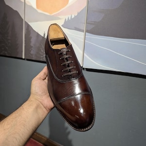 Peut inclure: Une chaussure Oxford en cuir marron avec un bout rapporté, présentant une finition polie et des lacets marron foncé. La chaussure est tenue dans une main, mettant en valeur son design élégant et son savoir-faire. Cette chaussure classique est adaptée aux occasions formelles.