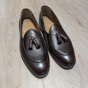 May include: A pair of dark brown leather loafers with a textured finish. Each shoe features a rounded vamp with a tassel detail. The shoes are set on a light brown wooden floor.