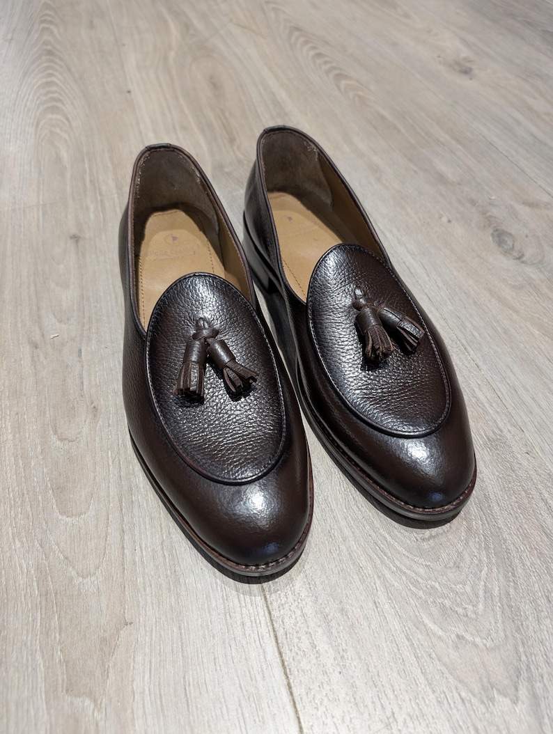 May include: A pair of dark brown leather loafers with a textured finish. Each shoe features a rounded vamp with a tassel detail. The shoes are set on a light brown wooden floor.