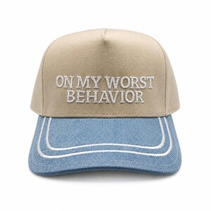 May include: A beige baseball cap featuring the white embroidered text ON MY WORST BEHAVIOR on the front panel. The cap has a light blue denim brim with two parallel white stitched lines following the curved edge of the visor.