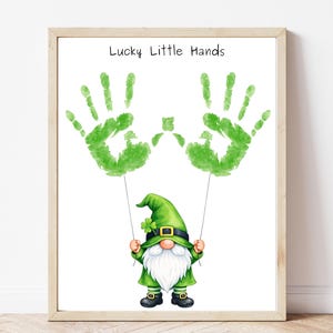 Könnte beinhalten: Gerahmter Druck mit grünen Handabdrücken und einem Cartoon-Gnom. Der Gnom, in grüner Kleidung und Hut, hält die Handabdrücke an Schnüren. Der Text "Lucky Little Hands" befindet sich darüber. Das Bild ist in einem Holzrahmen.