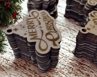 Puede incluir: Montones de adornos de árbol de Navidad de madera con la inscripción "MERRY CHRISTMAS". Los adornos son de color marrón claro con un borde marrón oscuro. Se ve una ramita de hoja perenne con bayas rojas.