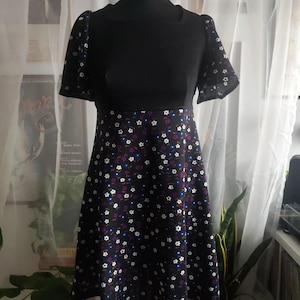 Puede incluir: Vestido negro con mangas cortas y escote cuadrado. El corpiño es negro liso, mientras que la falda presenta un estampado floral con flores blancas, moradas y azules sobre fondo negro. El vestido se exhibe en un maniquí.