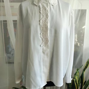 Peut inclure: Une blouse blanche avec un panneau de dentelle décoratif sur le devant et autour du col. La chemise à manches longues est présentée sur un mannequin. Le col est de forme arrondie avec une fermeture boutonnée.