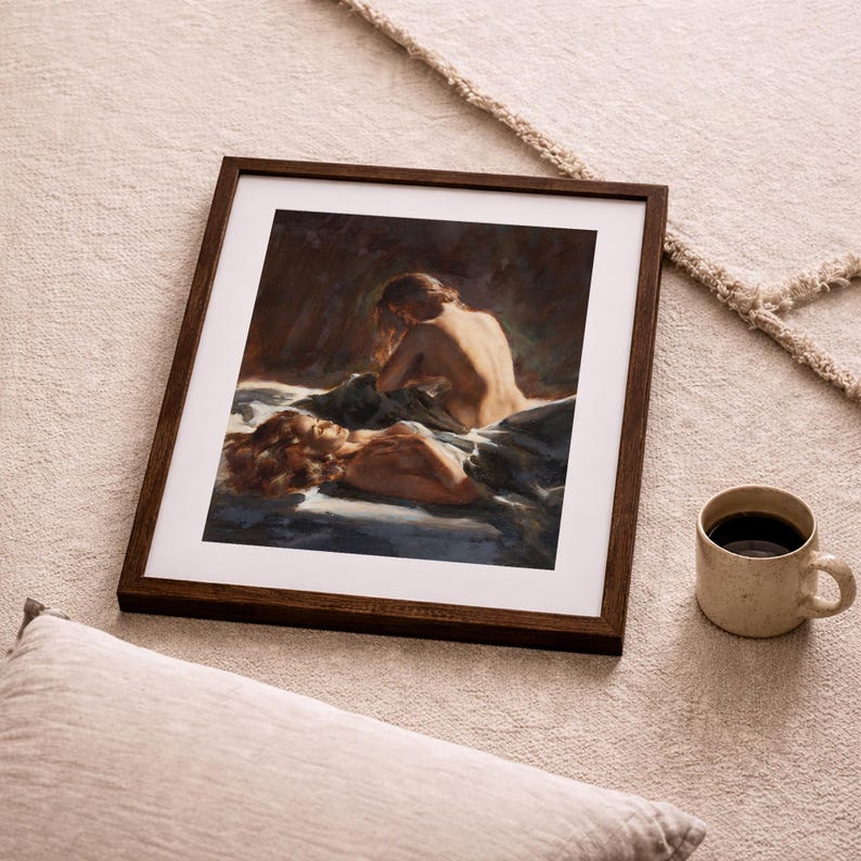 May include: A framed art print featuring a painting of two figures in a bed, with a dark brown wooden frame. The painting has a warm color palette with shades of brown, beige, and blue. A beige mug with coffee sits to the right of the frame.