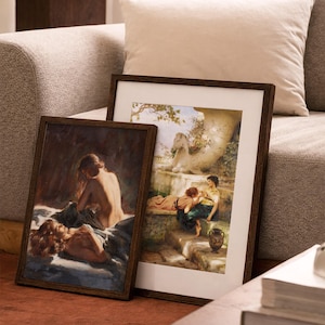 May include: Two framed art prints with dark wood frames. One print depicts a reclining figure, the other shows figures in a garden setting. The prints are leaning against a sofa with a neutral-colored cushion.
