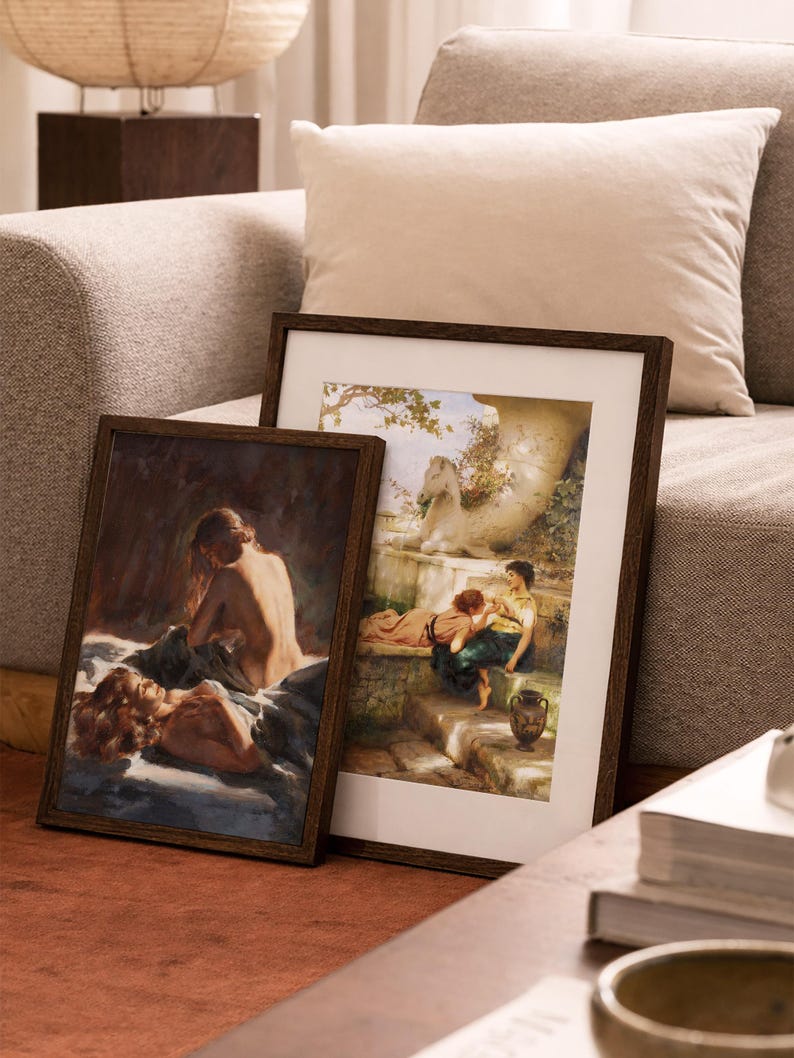May include: Two framed art prints with dark wood frames. One print depicts a reclining figure, the other shows figures in a garden setting. The prints are leaning against a sofa with a neutral-colored cushion.