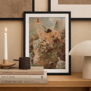 May include: A framed art print featuring a painting of winged figures amidst lush flowers and a swan in a pond. The artwork is displayed in a black frame, resting on a stack of three books next to a white candle and a white mushroom-shaped lamp.