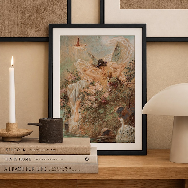 May include: A framed art print featuring a painting of winged figures amidst lush flowers and a swan in a pond. The artwork is displayed in a black frame, resting on a stack of three books next to a white candle and a white mushroom-shaped lamp.