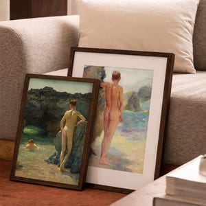 Op de afbeelding: Twee ingelijste kunstdrukken met klassieke schilderijen van naakte figuren aan zee. De prints hebben donkerbruine houten frames en worden tentoongesteld op een roodbruin tapijt voor een beige bank.