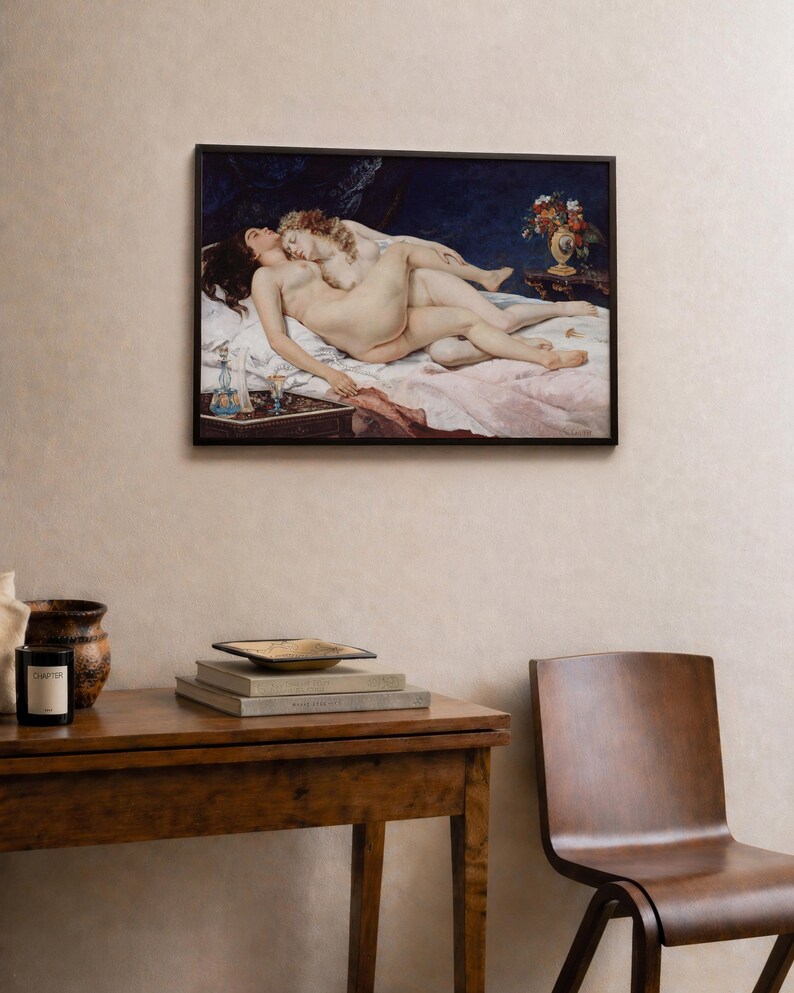 May include: A framed horizontal wall art print depicting two figures reclining on a bed against a dark blue background. The scene includes a small table with a vase of flowers and a glass decanter. The figures are positioned on white and pink linens.