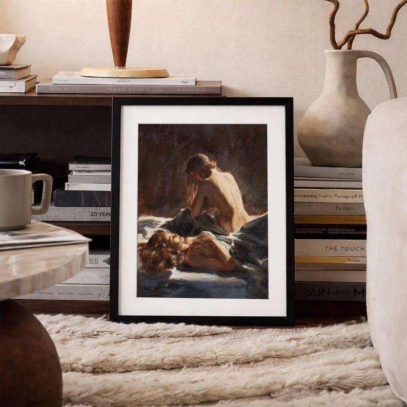 May include: A framed artwork featuring a painting of two figures in a dimly lit setting. One figure is seated, and the other is lying down. The artwork is displayed on a shelf with books and a decorative lamp.